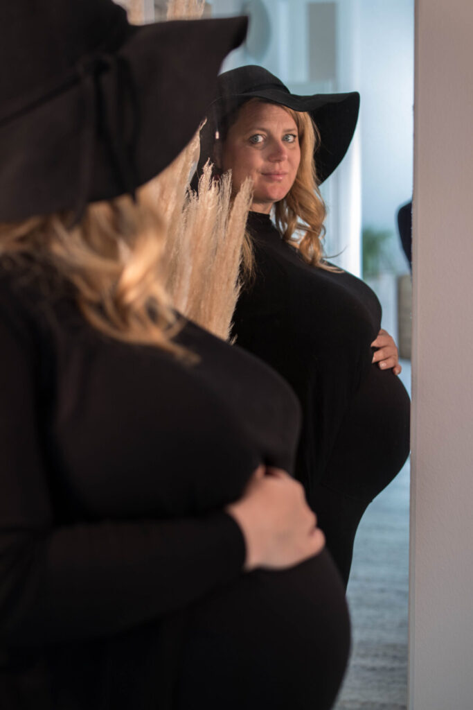 Schwangere Frau mit schwarzem Hut, die ihren Bauch in einem Spiegel betrachtet, umgeben von Trockenblumen.