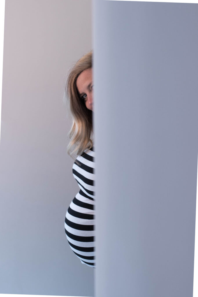 Eine schwangere Frau mit blonden Haaren in einem gestreiften Kleid steht teilweise hinter einer Wand.